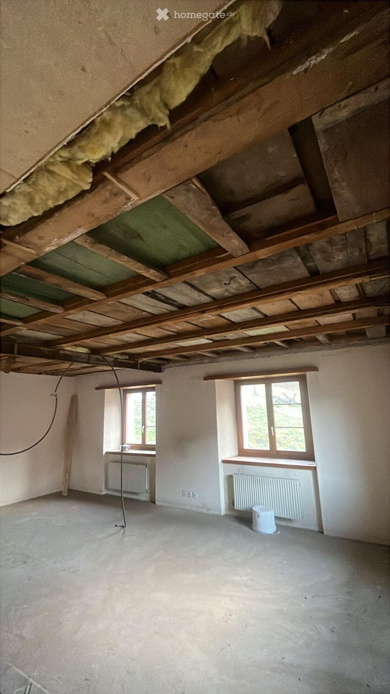 Empty room with exposed wooden beams and insulation in the ceiling, two windows, radiators, and a white bucket on the floor.