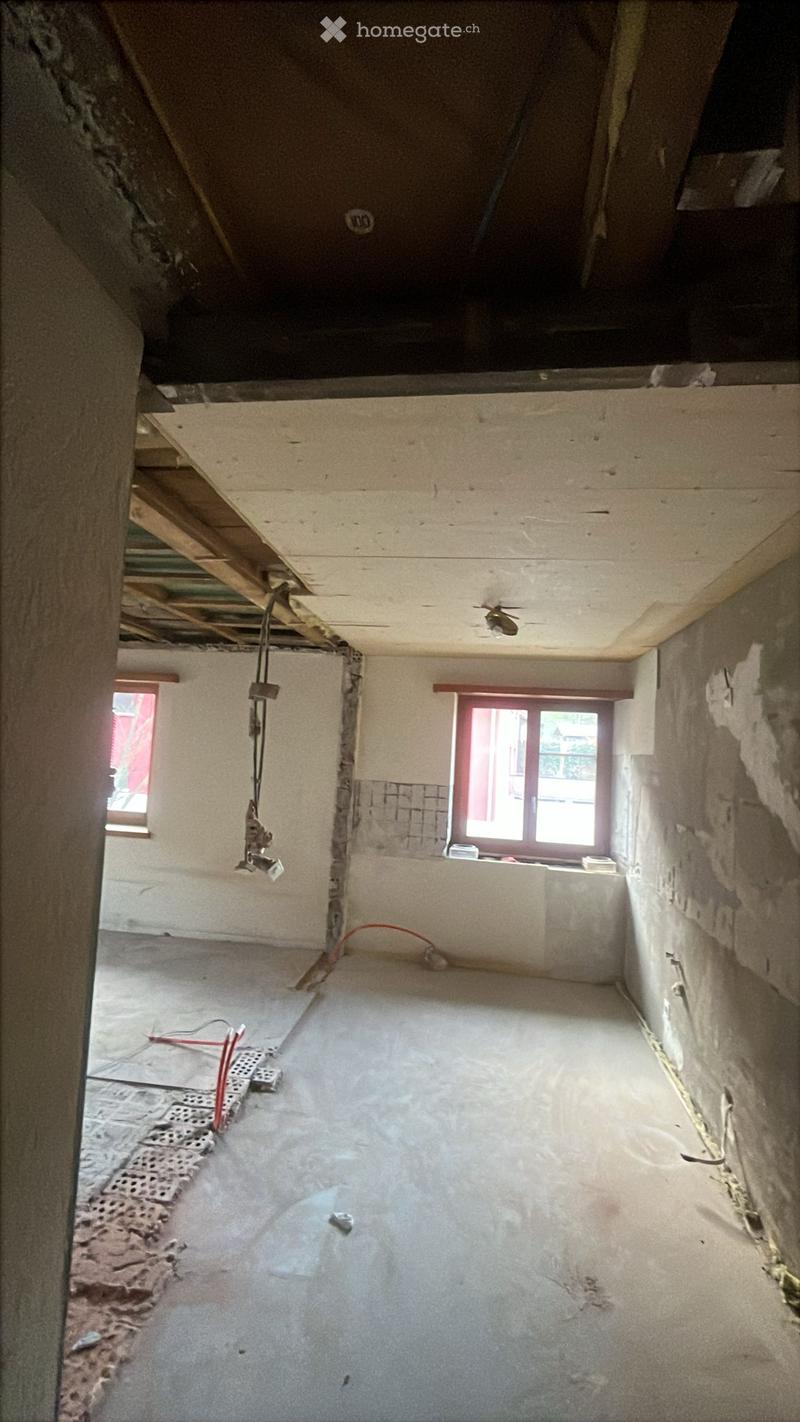 Empty room with unfinished concrete floor and exposed wooden beams on the ceiling. Walls are partially plastered, with visible electrical wiring and a window with red frame.