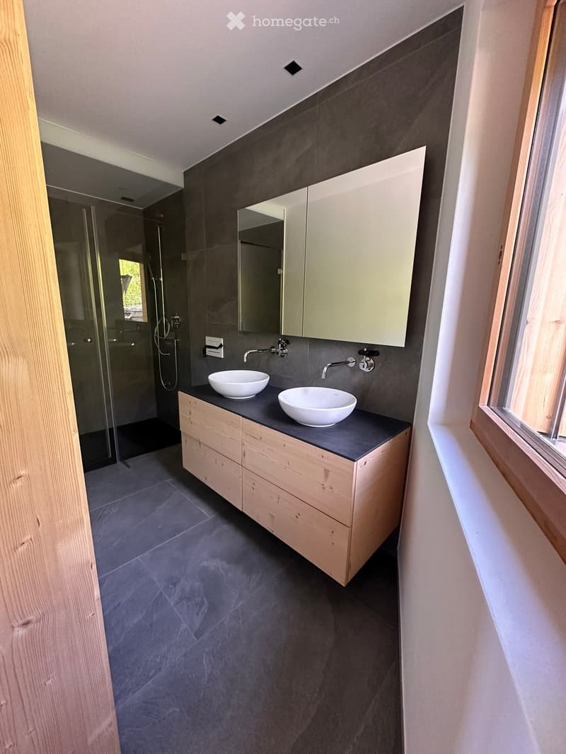 Modern bathroom with two white sinks on a wooden vanity, dark grey tiles, and a large mirrored cabinet above.