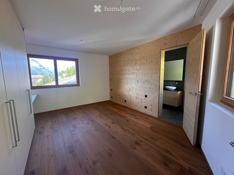 Empty room with wooden flooring, white walls, and a large window overlooking a mountain view. An adjacent bathroom with a sink and mirror is visible through an open doorway.