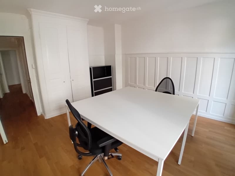 A modern, minimalist office space with a white table, black office chair, and a small black cabinet. The room features light wooden flooring and white walls with paneling.