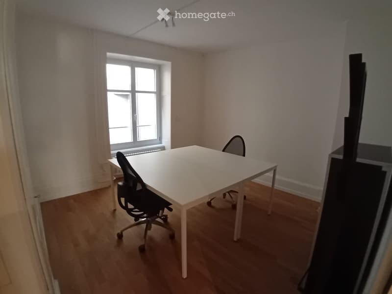 A small, unfurnished room with a white table and a single black chair, wooden flooring, and a window with curtains.