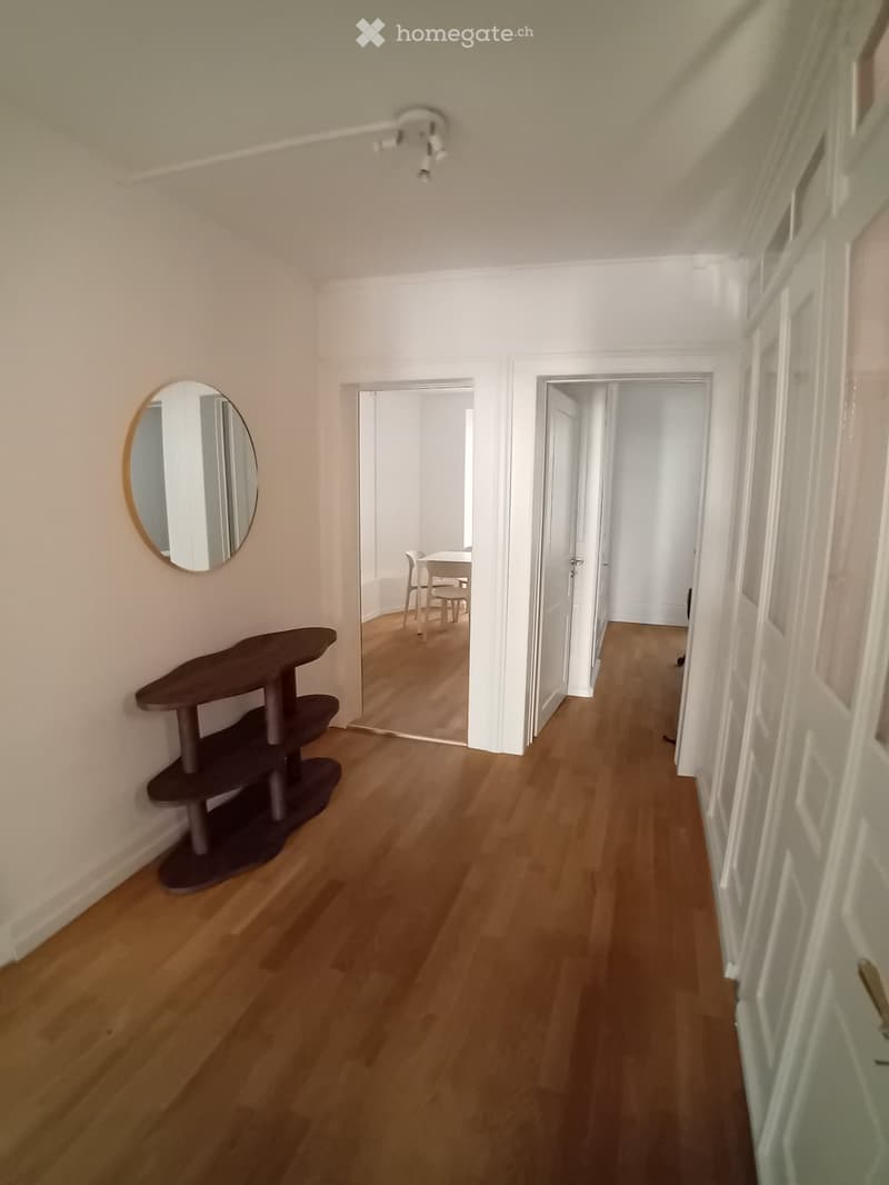 A narrow hallway with wooden flooring, white walls, and a small shelf and mirror on the left side. Two open doors lead to adjacent rooms.