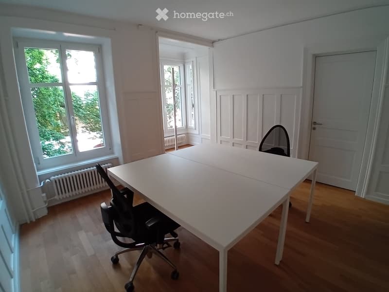 Bright room with wooden flooring, large windows, white table, and black office chair.