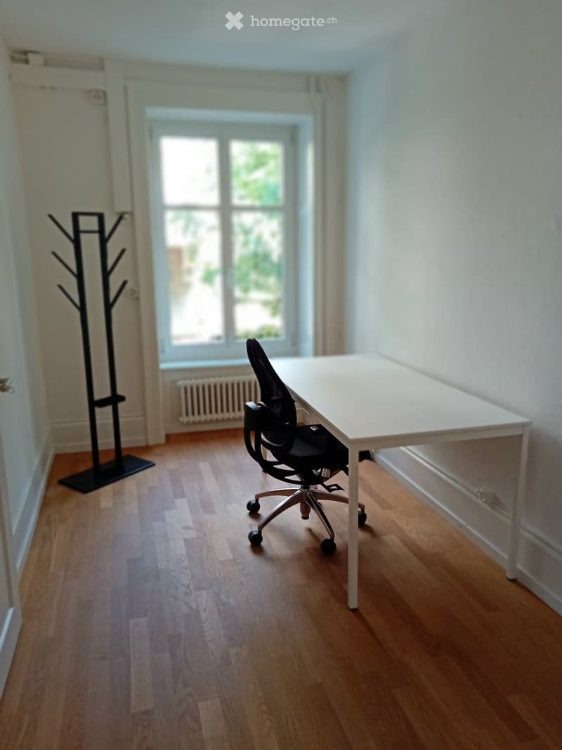 A small, well-lit room with wooden flooring, a white table, a black office chair, and a coat rack near the door.