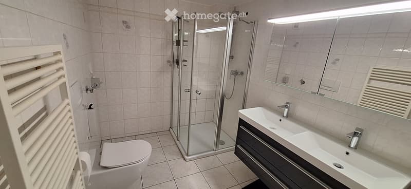 Modern bathroom with a white tiled shower stall, a white toilet, a double sink with a mirrored cabinet, and a white radiator on the wall.