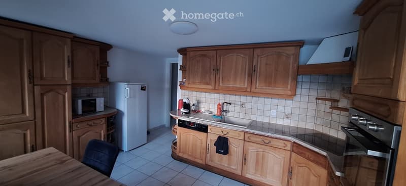 Well-lit kitchen with wooden cabinets, a refrigerator, microwave, sink, oven, and dishwasher. The countertops are tiled, and there is a small table and chair in the foreground.