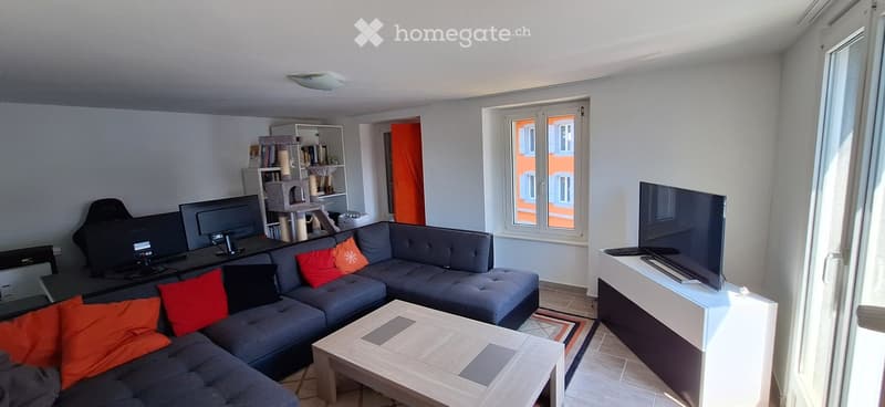 Modern living room with a grey sectional sofa, colorful cushions, a wooden coffee table, a TV stand, a cat tree, and a bookshelf. Large windows provide natural light.