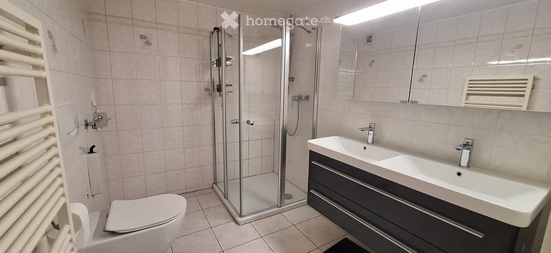 Modern bathroom with a white tiled shower stall, a double sink with a modern cabinet, a toilet, and a radiator. The walls are tiled in light beige tiles, and the floor is tiled in a darker shade.