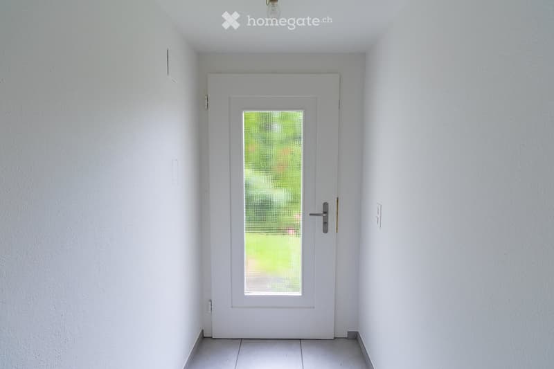 A clean, white-walled hallway with a central white door featuring a frosted glass panel. The floor is tiled, and the ceiling has a simple light fixture.