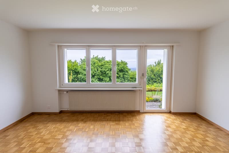 Empty room with wooden flooring, white walls, large windows, and a sliding door leading to a garden area.