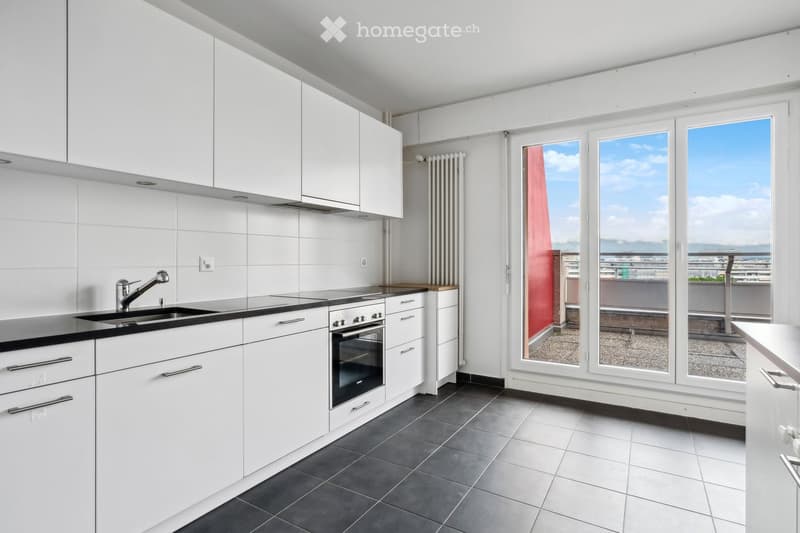 Modern kitchen with white cabinets, black countertops, and a sink. Large windows offer a view of the city and a balcony area.