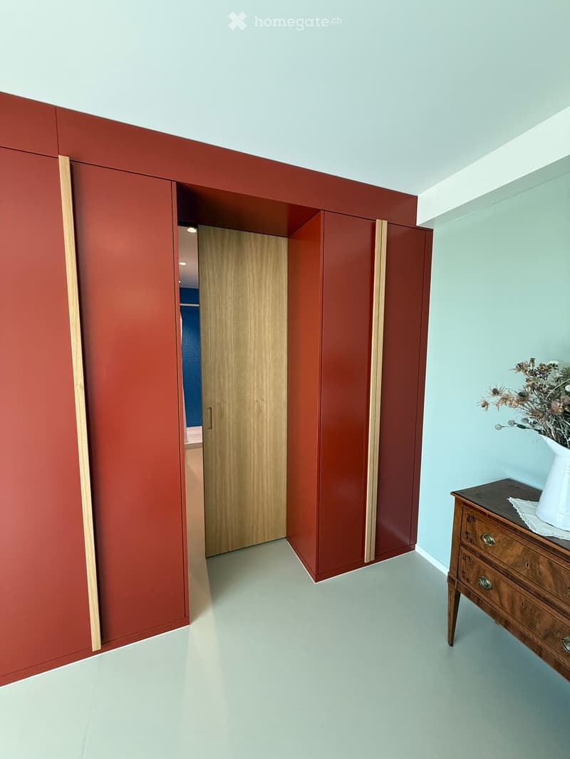 Modern hallway with red accent wall, light blue walls, white floor, wooden door, chest of drawers with vase.