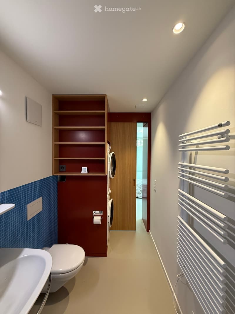 Modern bathroom with a bathtub, toilet, radiator, built-in shelf, and a washing machine in the background.