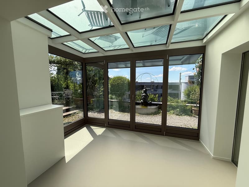 Bright, open room with large glass doors and skylights, leading to a garden area with a fountain and outdoor seating.