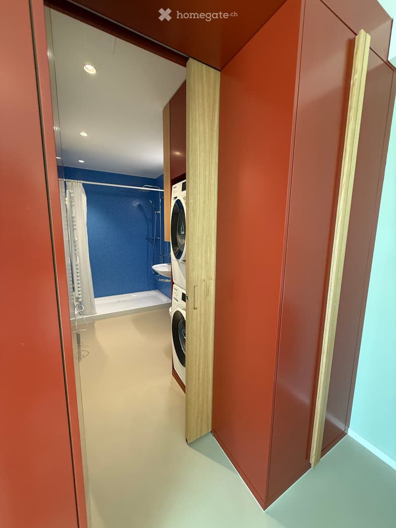 Modern bathroom with blue tiled shower area, white bathtub, washing machine, and sleek wooden cabinetry.