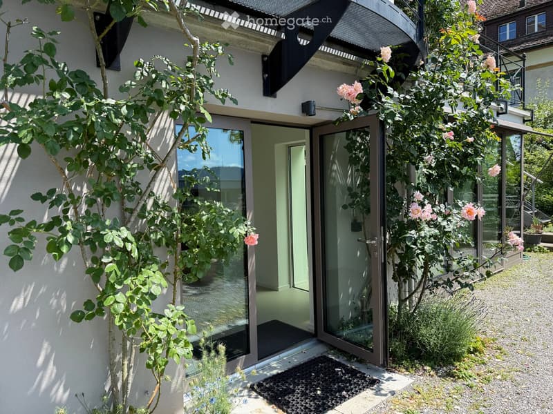 A modern house entrance with large glass doors, surrounded by lush greenery and climbing roses. The pathway is gravel, and the entrance features a doormat.