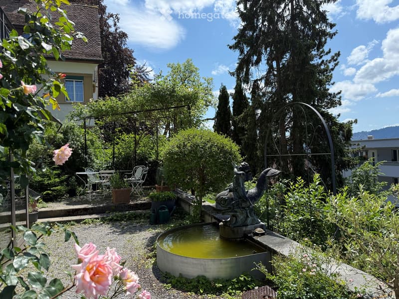 A lush garden with a central fountain, surrounded by greenery, potted plants, and outdoor furniture including chairs and a table. The area is enclosed by trees and features a decorative statue near the fountain.