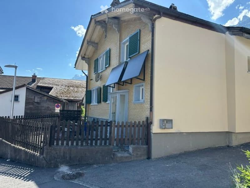 Two-story house with beige walls, green shutters, and a wooden fence in front. The house features a small balcony with a shuttered window and a covered entrance area.