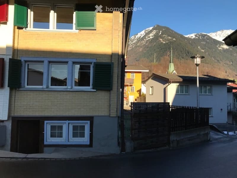 Two-story building with yellow facade, white-framed windows, green shutters, and a view of snow-capped mountains in the background.