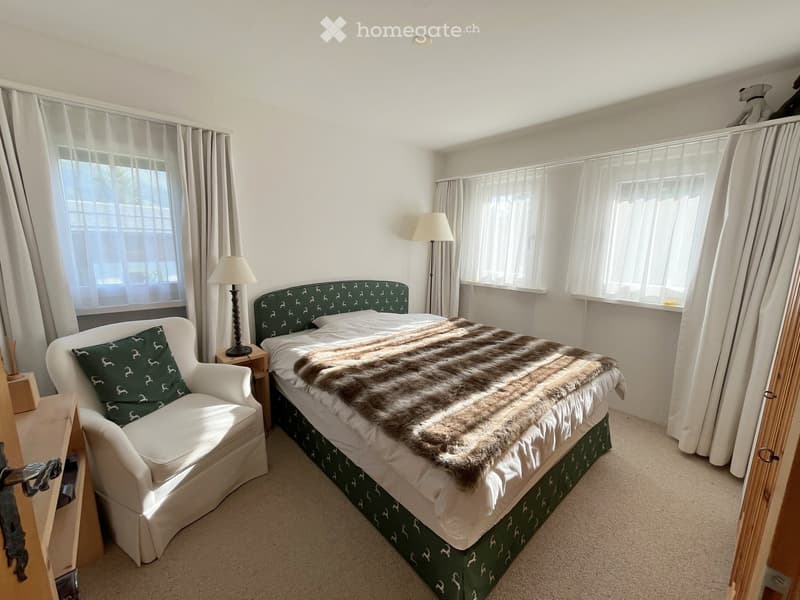 Cozy bedroom with a large bed, fur throw, white armchair, and lamps. The room features large windows with white curtains and a neutral color palette.