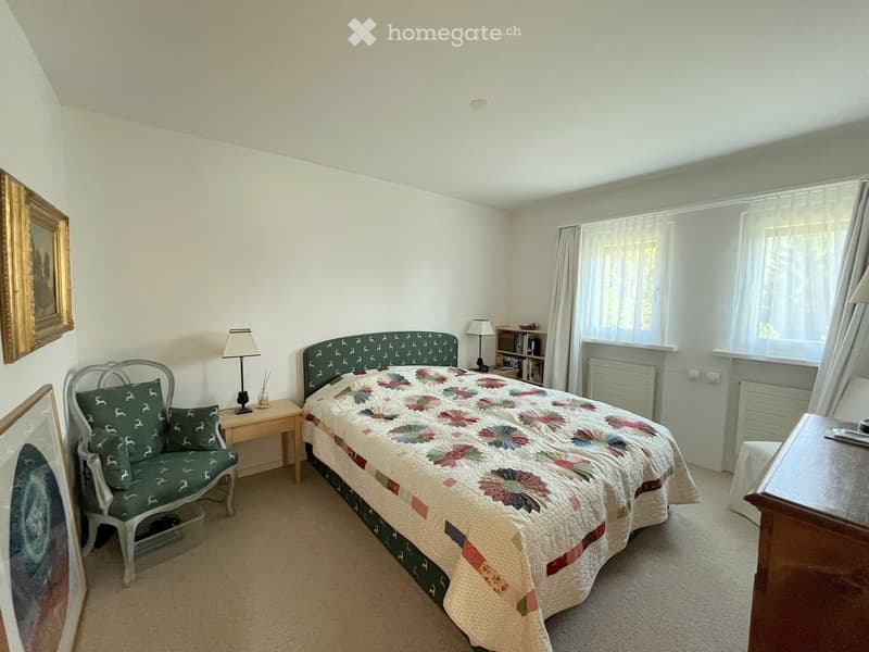 Cozy bedroom with a quilted bed, a green armchair, a wooden nightstand, and a lamp. The room features large windows with curtains and a radiator below them.