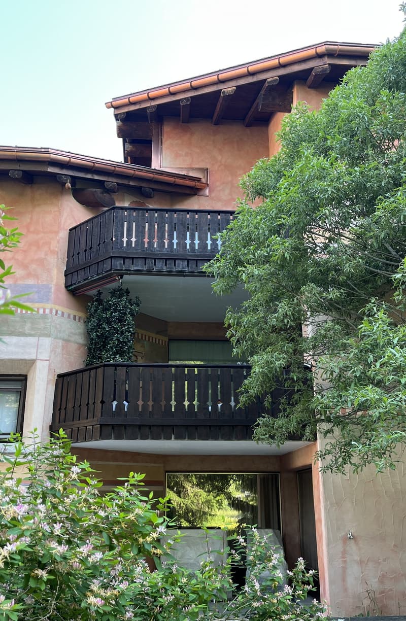 Two-story building with wooden balconies, terracotta roof, and surrounding greenery.