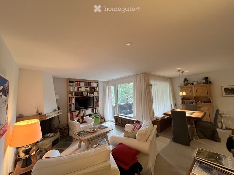 Bright living room with large windows and a view, featuring a fireplace, sofa, chairs, coffee table, bookshelf with TV, and a small desk area.