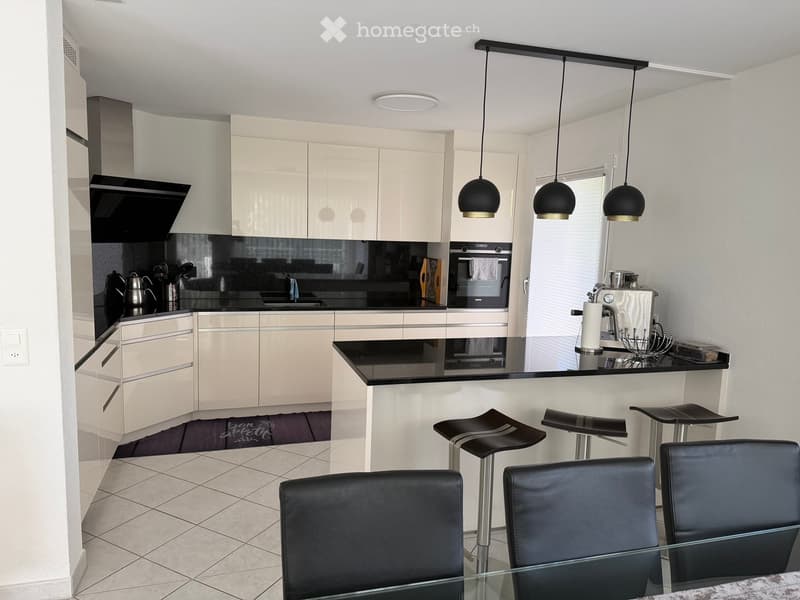 Modern kitchen with white cabinets, black countertops, and a central island with bar stools. Features include a coffee maker, sink, and oven.