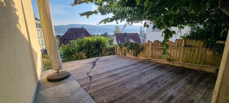 Wooden deck terrace with a view of a lake and distant mountains, surrounded by greenery and a wooden fence.