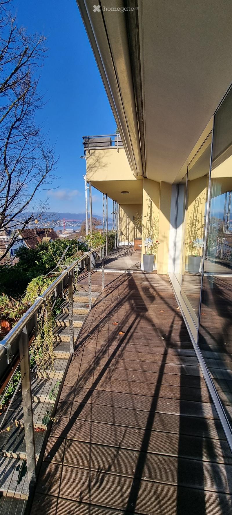 Wooden balcony with metal railing, potted plants, and a view of the city and water.