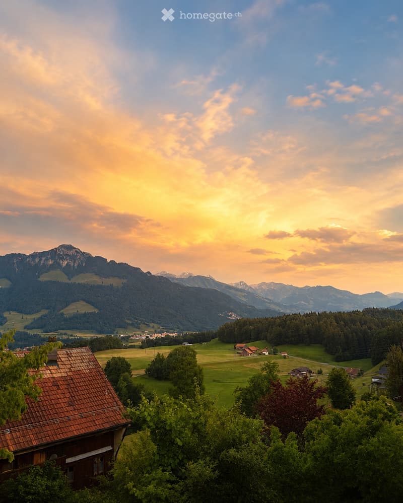 Scenic view of a rural landscape with a wooden house in the foreground, green fields, scattered trees, and distant mountains under a sunset sky.