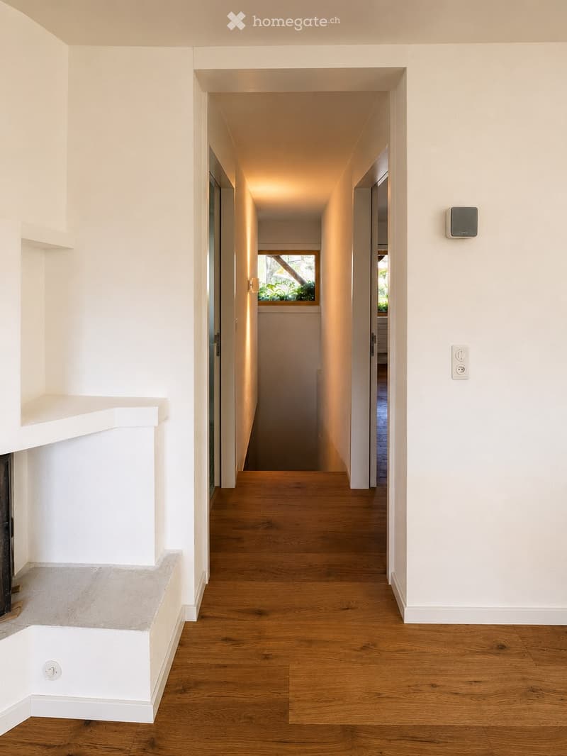 Modern hallway with light wooden flooring, white walls, recessed lighting, and a small window at the far end.