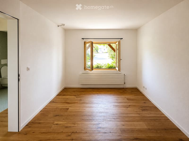 Empty room with wooden flooring, white walls, and a large window with wooden frames. A radiator is placed below the window, and a toilet is visible in an adjacent room through an open doorway.
