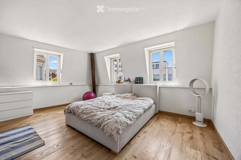 Bright bedroom with wooden flooring, large windows, a bed with a patterned duvet, a nightstand, a fan, and a colorful ball on the floor.