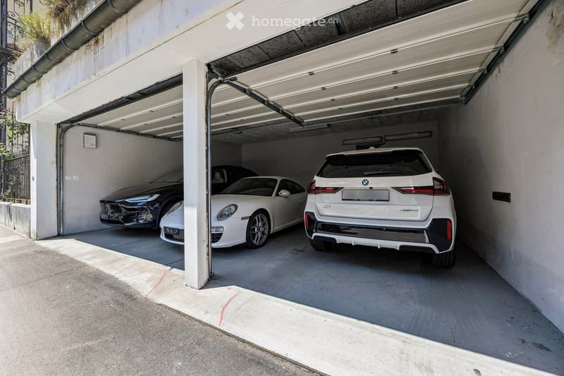 Underground parking area with three cars parked inside, including a white BMW and a white Porsche, under a concrete ceiling with fluorescent lighting.