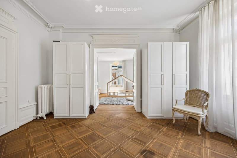 Elegant hallway with wooden herringbone flooring, white walls, large built-in wardrobes, a classic white chair, and a radiator. A view into an adjacent room shows a crib and soft rug.