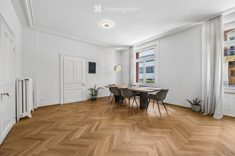 Bright dining area with herringbone wooden flooring, large window with white curtains, modern dining table with six chairs, two potted plants, radiator, and a floor lamp.