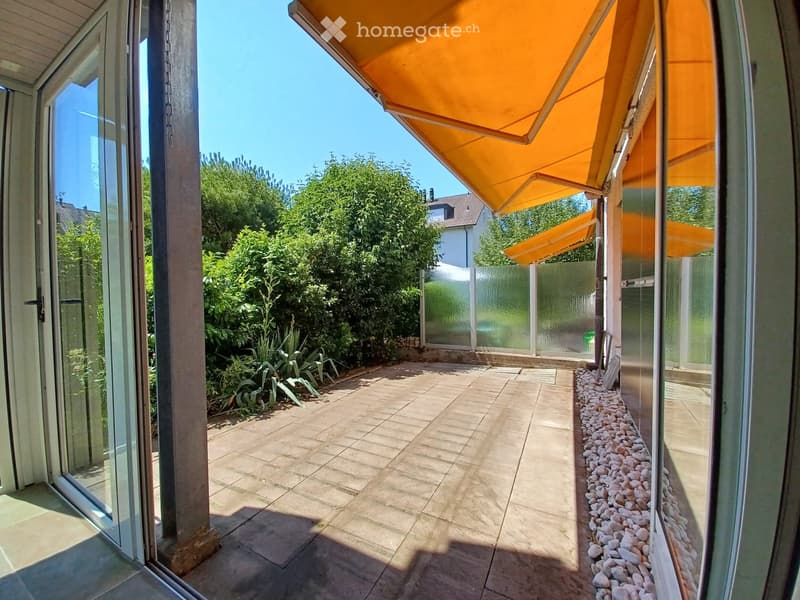 A covered patio with tiled flooring, glass walls, and a view of a garden with greenery and a neighboring house.