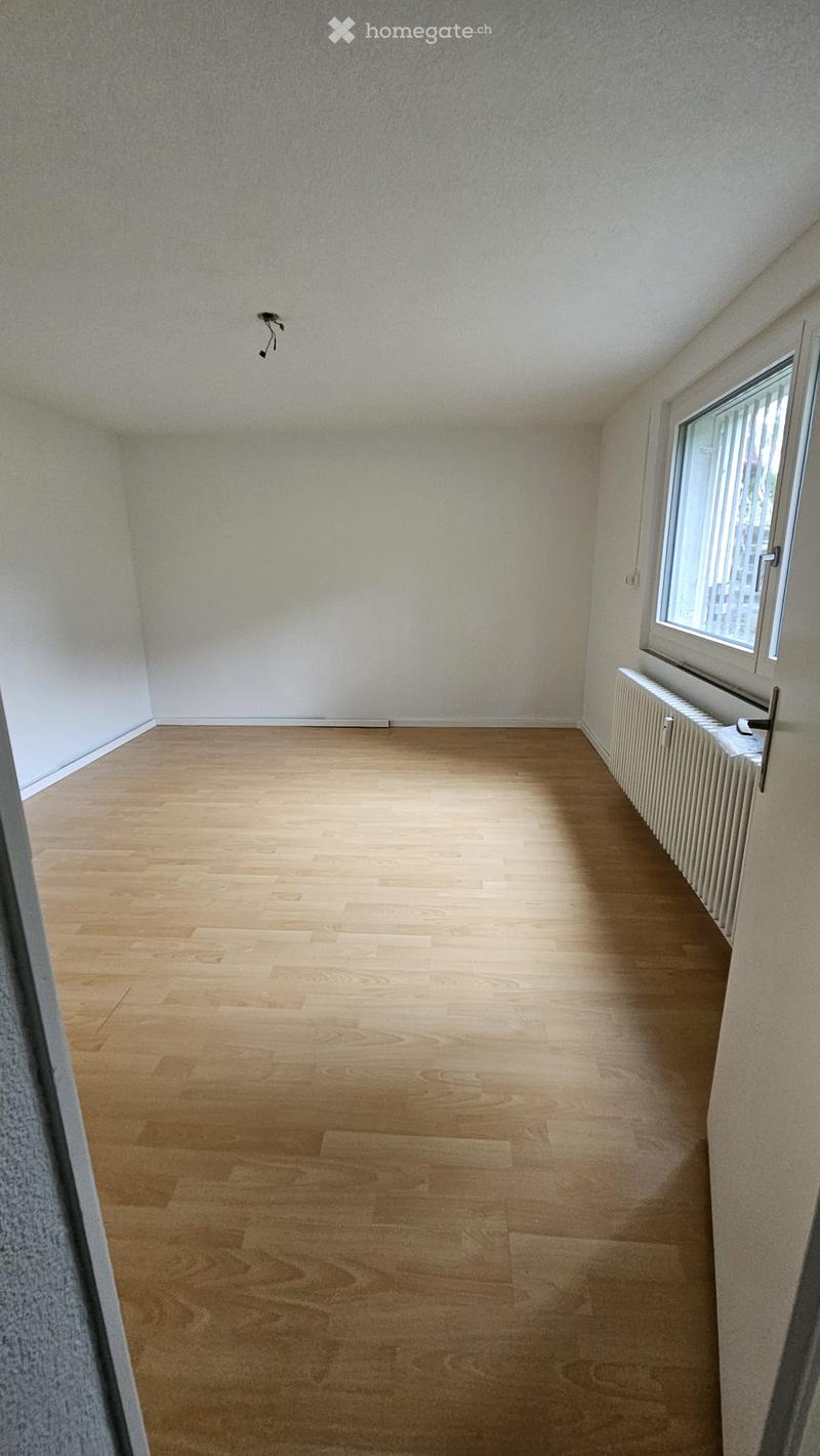 Empty room with light wooden flooring, white walls, a single window with blinds, and a radiator below the window.