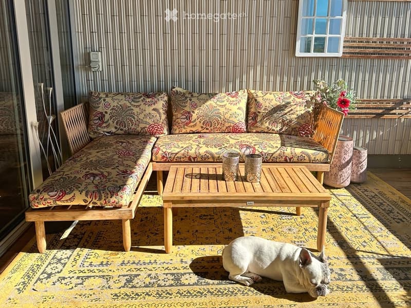 Balcony with a wooden sofa, wooden coffee table, patterned cushions, and a white dog lying on a yellow rug.