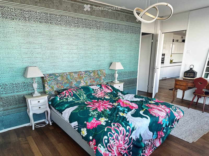 A modern bedroom with a large bed covered in a floral duvet, two nightstands with lamps, a wooden floor, and a view into an adjacent kitchen area.