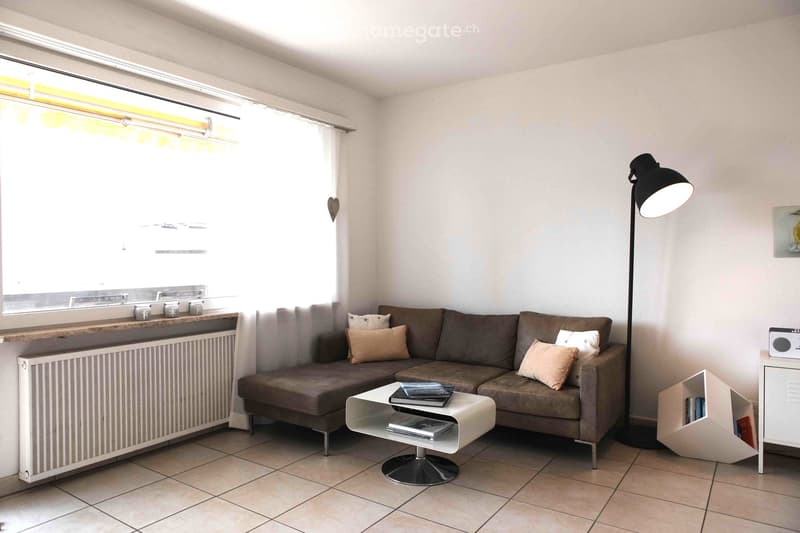 Modern living room with a grey sectional sofa, beige throw pillows, a white coffee table with books, a floor lamp, and a small shelf with books. The room features tiled flooring and large windows with blinds.