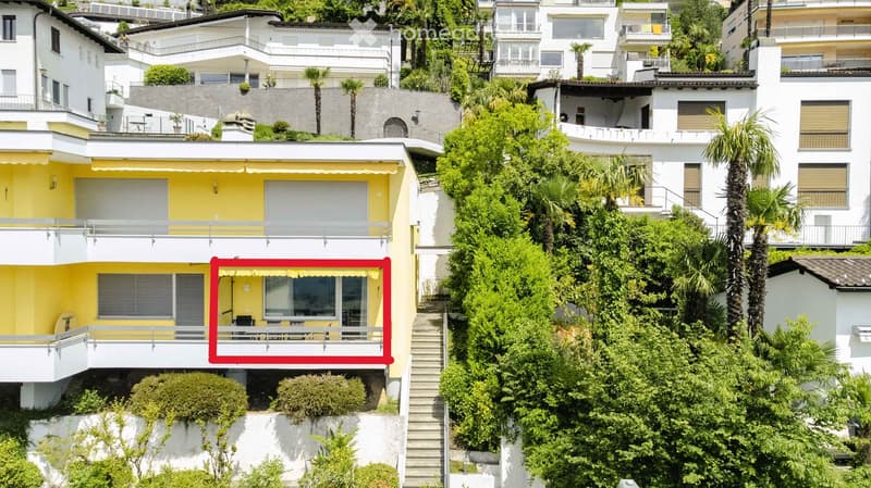 Balcony with a table and chairs, overlooking a lush, green hillside with neighboring houses and palm trees.