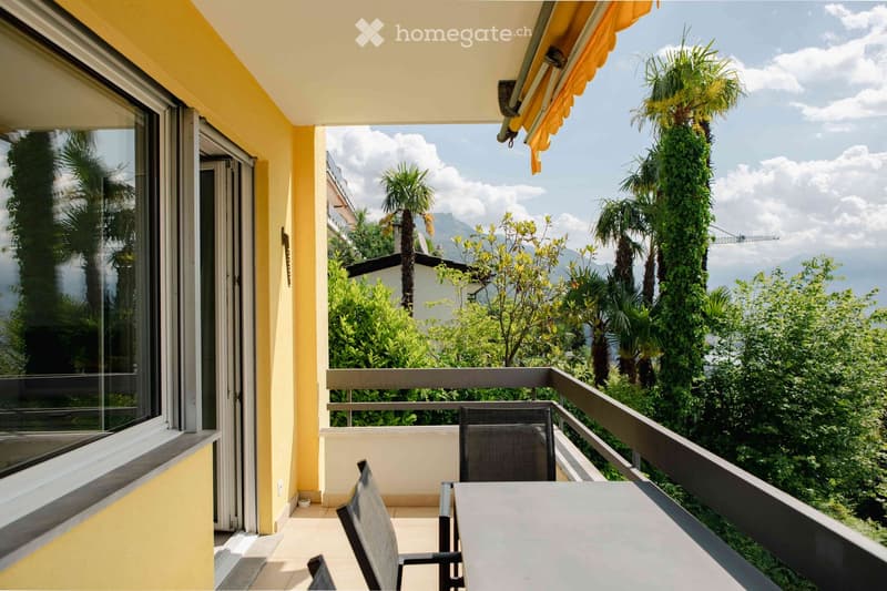 Balcony with a table and chairs, overlooking a lush green area with palm trees and mountains in the background.