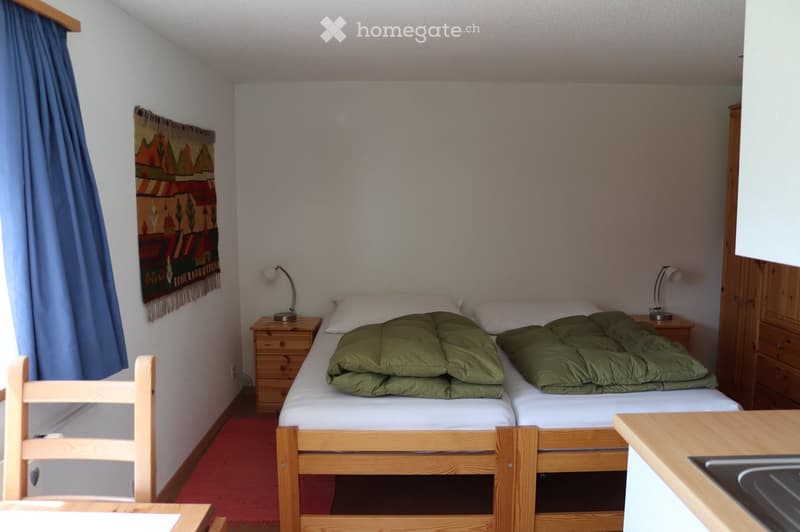 Cozy bedroom with a twin-sized bed, green duvet, two nightstands with lamps, a wooden chair, and a colorful tapestry on the wall.