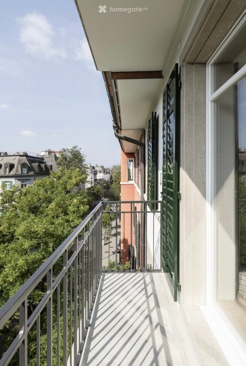 Balcony with metal railing, overlooking a street with trees and buildings.