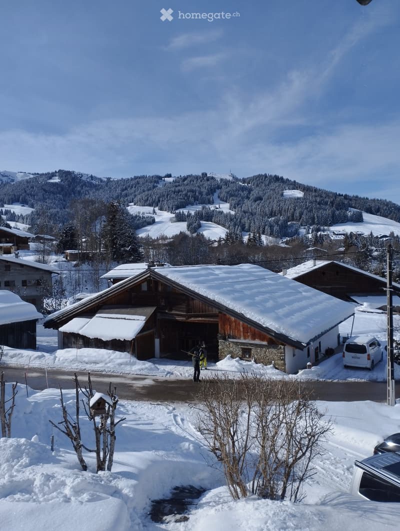 A large wooden house with a stone base, covered in snow, situated in a snowy mountain landscape with other houses and a clear blue sky.