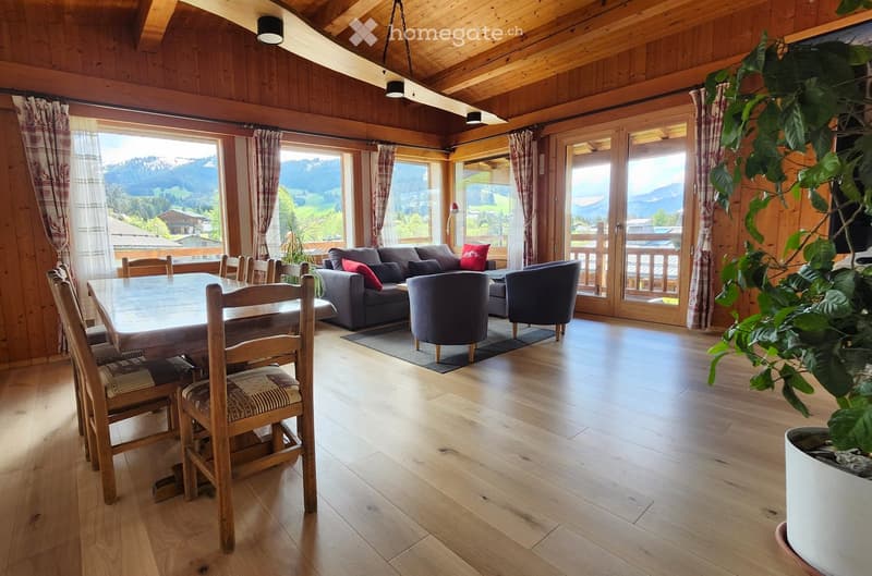 A spacious, well-lit living room with wooden floors, large windows offering a scenic mountain view, a grey sofa with red cushions, a wooden dining table with chairs, and a potted plant in the corner.