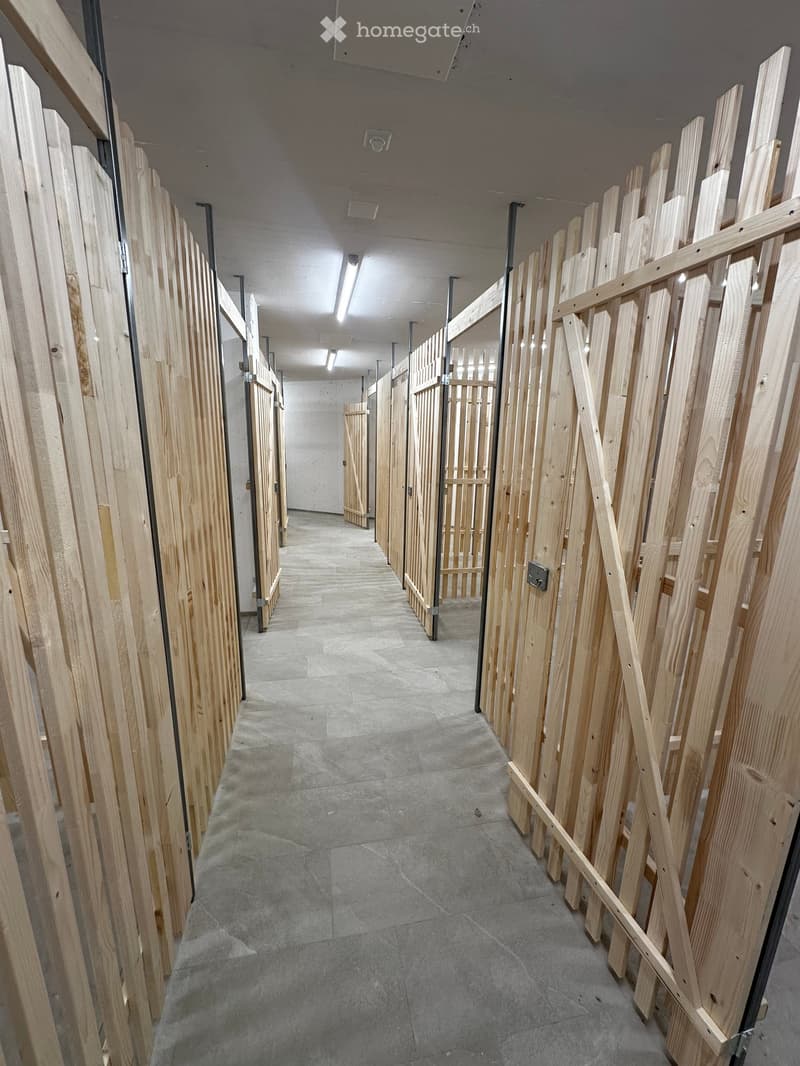 Empty hallway with wooden partitions on both sides, concrete floor, and fluorescent lighting.